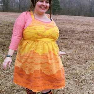 May include: A woman wearing a yellow and orange dress with a pink cardigan and black and white striped socks. She is standing in a field of dry grass.
