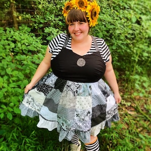 May include: A person wearing a black and white striped shirt with a black overall dress and a sunflower crown. The dress has a patchwork design with a ruffled hem. The person is standing in a grassy area.