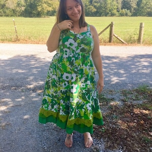 May include: A woman wearing a green and white floral maxi dress with a green lace trim. The dress has a fitted bodice and a flowing skirt. She is standing on a gravel path in front of a green field.