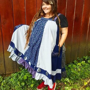 May include: A woman wearing a blue, white, and purple patchwork dress with a moon and star design. The dress has a ruffled hem and is worn over a black shirt. The woman has long brown dreadlocks and is standing in front of a red wooden fence.