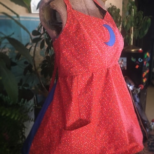 May include: Red and white floral print apron with a blue crescent moon patch and a large pocket.