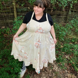 May include: A woman wearing a beige floral print dress with a ruffled hem and black and white striped socks. She is standing in a grassy area with a wooden fence in the background.