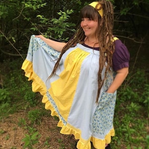 May include: A woman wearing a blue and yellow patchwork dress with a ruffled hem. She has long brown dreadlocks and is wearing a yellow headband.