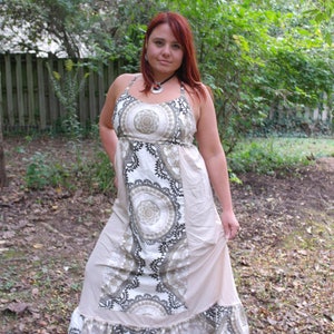 May include: A woman wearing a long, white maxi dress with a brown and white floral pattern. The dress has a ruffled hem and a halter neckline. She is standing in a wooded area with green trees and brown leaves on the ground.