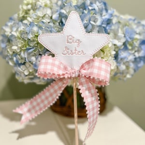 Op de afbeelding: Een witte stervormige decoratie met de woorden "Big Sister" in roze geborduurd. Het heeft een roze en witte gingham strik met pompon afwerking en een houten stok. De decoratie staat voor een boeket blauwe en witte hortensia's.