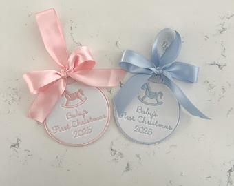Può includere: Due ornamenti natalizi ricamati, uno rosa e uno blu, ciascuno con un nastro di raso. Gli ornamenti presentano un disegno di cavallo a dondolo e il testo "Baby's First Christmas 2025".