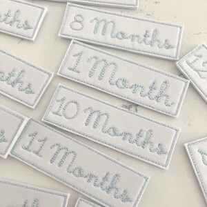 以下が含まれることがあります： 月を示す、薄い青色の刺繍が施された白い長方形の布パッチ。 パッチは薄い青色のステッチで縁取られています。 テキストには「1ヶ月」、「8ヶ月」、「10ヶ月」、「11ヶ月」が含まれています。