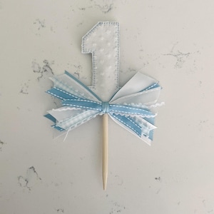 May include: A white fabric number one cake topper with a blue and white ribbon bow. The number one is made of white fabric with a dotted pattern and blue stitching. The bow has layers of white and blue ribbons, and a wooden stick.