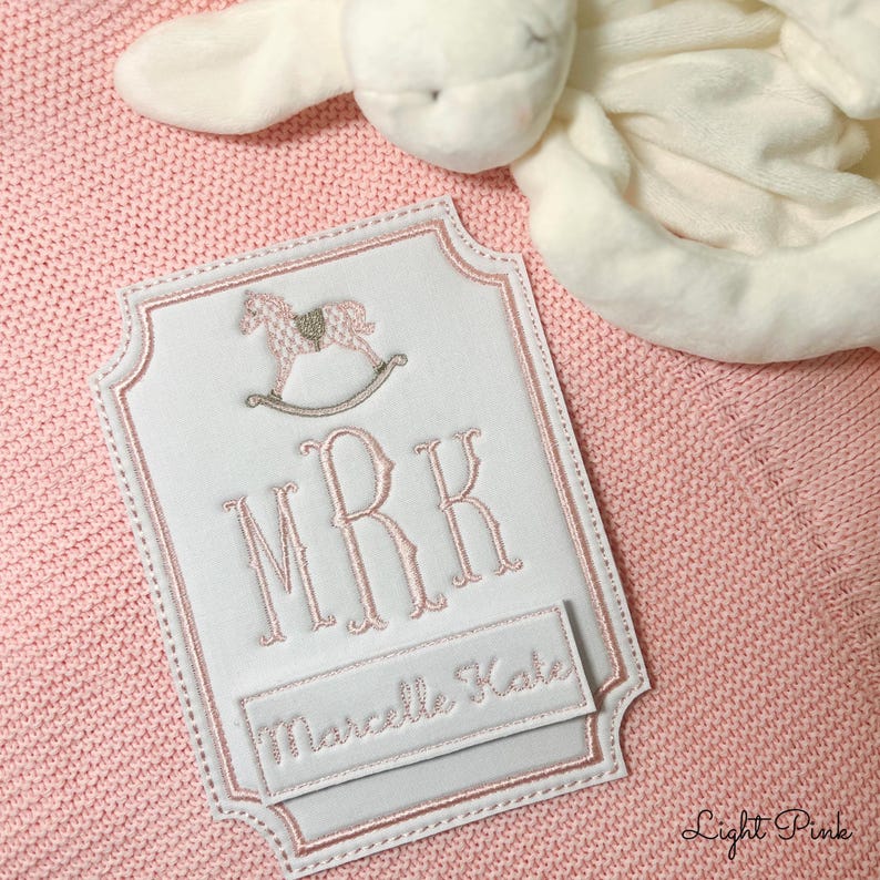 以下が含まれることがあります： イニシャル「MRK」と「Marcelle Kate」の名前が入った、白とピンクの刺繍入りベビーブランケットラベル。ロッキングホースのデザインで、ピンク色のニットブランケットの上に配置されています。「Light Pink」の文字が右下にあります。