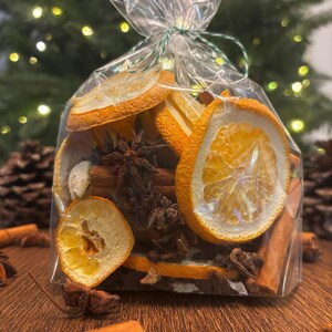 May include: A clear plastic bag filled with dried orange slices, cinnamon sticks, and star anise. The bag is tied with a green string and is sitting on a wooden surface.