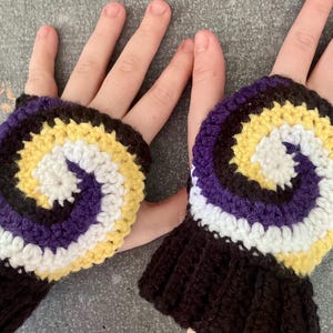 May include: Handmade crochet fingerless gloves in a non-binary pride flag design. The gloves feature a spiral pattern in white, yellow, purple, and black. The wrist cuffs are black.