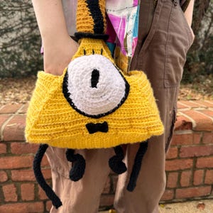 May include: A handmade yellow crochet bag shaped like a cartoon character with a large white eye, black pupil, and a small black bow tie. The bag has black legs and a black top hat. The bag is held in front of a person.