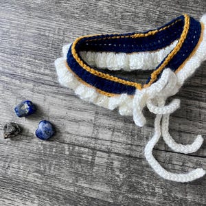 May include: A crocheted collar featuring a dark blue band with a thin mustard yellow border and a white ruffled edge. The collar includes two long white crocheted ties at the front. Three small, polished blue and grey stones sit nearby.