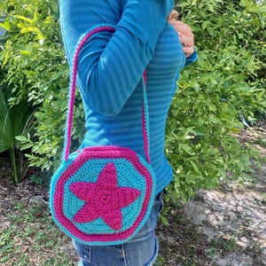 Op de afbeelding: Een handgemaakte, gehaakte crossbodytas. De tas heeft een turquoise body met een roze ster en rand. De band is roze en turquoise. De tas is rond met een achthoekige vorm. De tas wordt over de schouder gedragen.