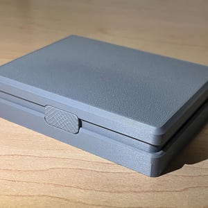 以下が含まれることがあります： グレーのプラスチック製の箱で、ヒンジ付きの蓋とテクスチャ加工されたラッチが付いています。箱は閉じています。
