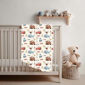 May include: A cream-coloured baby blanket featuring a repeating pattern of cartoon race cars, traffic cones, chequered flags, and street lamps. The design includes red, blue, and brown vehicles with smiling faces, arranged on a light background.
