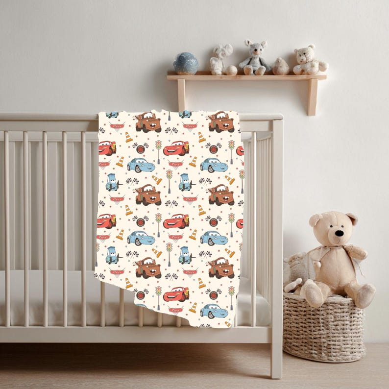 May include: A cream-coloured baby blanket featuring a repeating pattern of cartoon race cars, traffic cones, chequered flags, and street lamps. The design includes red, blue, and brown vehicles with smiling faces, arranged on a light background.