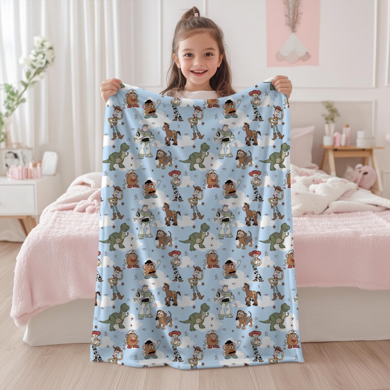 K&ouml;nnte beinhalten: Eine hellblaue Fleecedecke mit einem sich wiederholenden Muster aus Toy Story-Figuren wie Woody, Buzz Lightyear, Rex, Slinky Dog und Mr. Potato Head, durchsetzt mit kleinen wei&szlig;en Wolken und grauen Sternen auf der gesamten Stoffoberfl&auml;che.