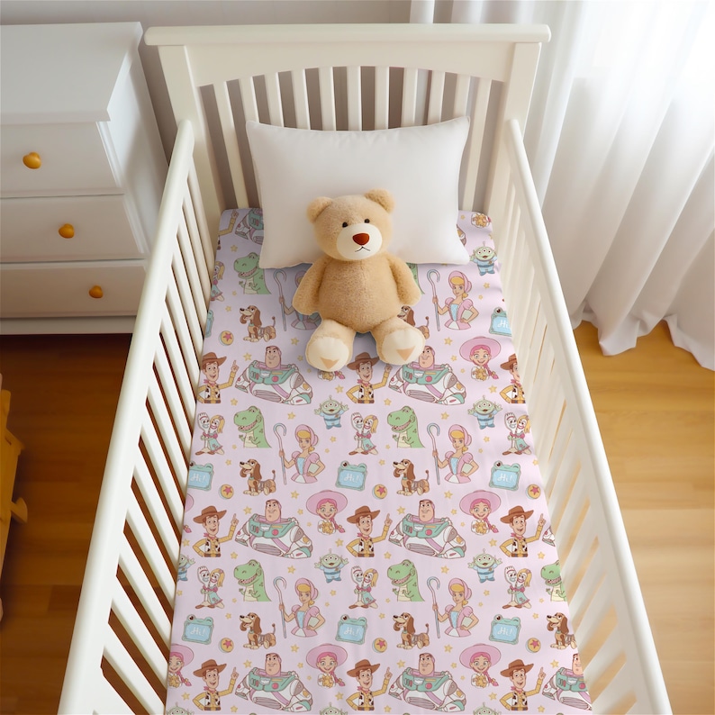 May include: A light pink crib sheet featuring a repeating pattern of Toy Story characters including Woody, Buzz Lightyear, Bo Peep, Rex, Slinky Dog, and Alien, with a light brown plush teddy bear sitting in the center of the crib mattress.