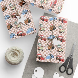 May include: Wrapping paper featuring a repeating pattern of cartoon race cars and tow trucks wearing Santa hats and scarves, set against a light pink and white checkered background with small scattered stars, tied with brown twine and white tags.