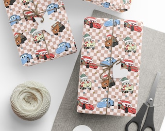 May include: Wrapping paper featuring a repeating pattern of cartoon race cars and tow trucks wearing Santa hats and scarves, set against a light pink and white checkered background with small scattered stars, tied with brown twine and white tags.