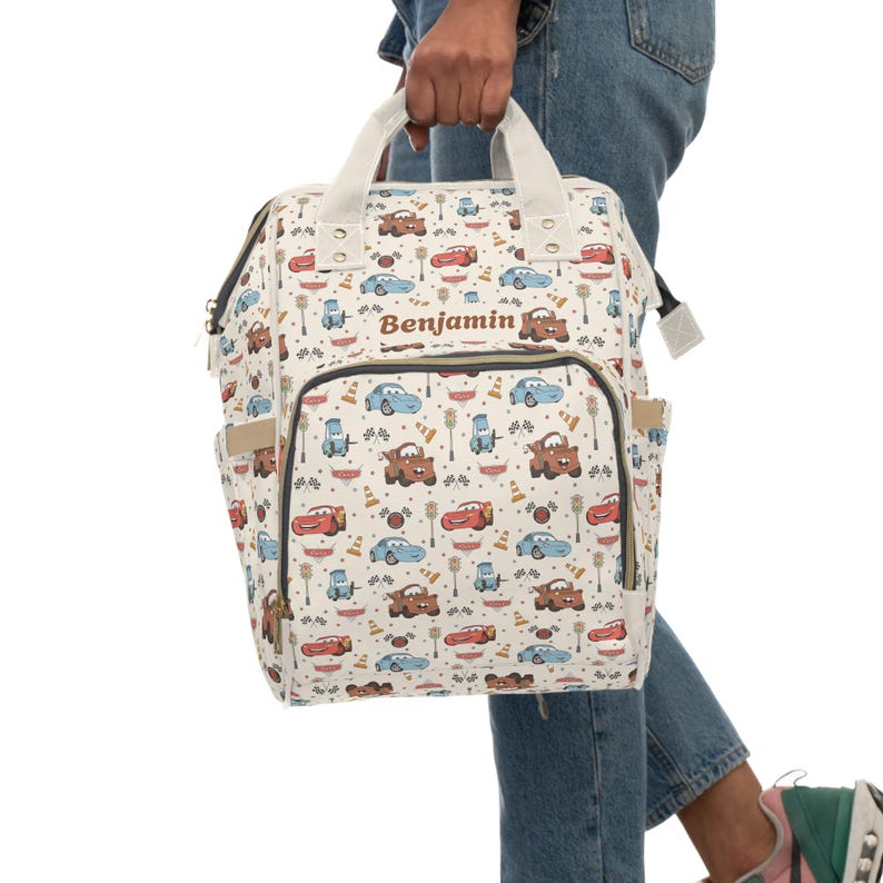 Puede incluir: Una mochila de color crema con un patr&oacute;n repetitivo de coches de carreras de dibujos animados, conos de tr&aacute;fico y banderas a cuadros. La bolsa tiene dos asas superiores, un bolsillo frontal con cremallera y el nombre Benjamin en marr&oacute;n.