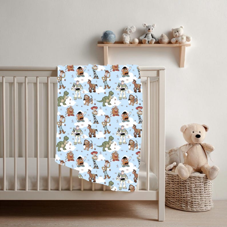 K&ouml;nnte beinhalten: Eine hellblaue Babydecke mit einem sich wiederholenden Muster aus verschiedenen Zeichentrickfiguren der Toy Story-Reihe, darunter ein Weltraum-Ranger, ein Cowboy, ein Cowgirl, ein Dinosaurier, eine Kartoffelfigur und ein Dackel, durchsetzt mit wei&szlig;en Wolken und Sternen.