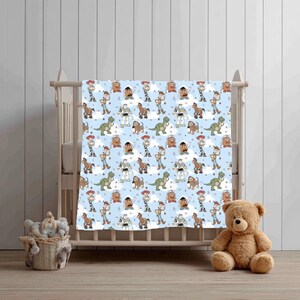 K&ouml;nnte beinhalten: Eine hellblaue Babydecke mit einem sich wiederholenden Muster aus Zeichentrickfiguren, darunter ein Cowboy, ein Weltraum-Ranger, ein Cowgirl, ein Dinosaurier, eine Kartoffelfigur, ein Pferd und ein Hund, durchsetzt mit kleinen wei&szlig;en Wolken.