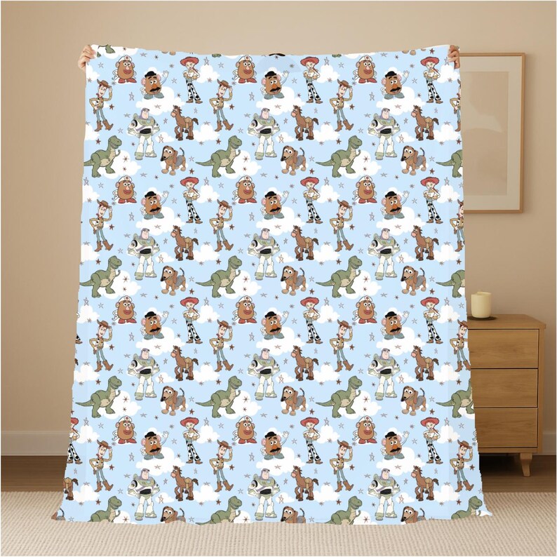 K&ouml;nnte beinhalten: Eine hellblaue Fleecedecke mit einem sich wiederholenden Muster aus Toy Story-Figuren wie Woody, Buzz Lightyear, Jessie, Rex, Mr. Kartoffelkopf, Slinky Dog und Bullseye, durchsetzt mit kleinen wei&szlig;en Wolken und braunen Sternformen.