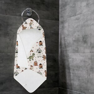 May include: A hooded baby towel with a pattern of cartoon characters, including a cowboy, a space ranger, and a green dinosaur, hangs on a silver towel ring. The towel is white on the inside and has a beige background.