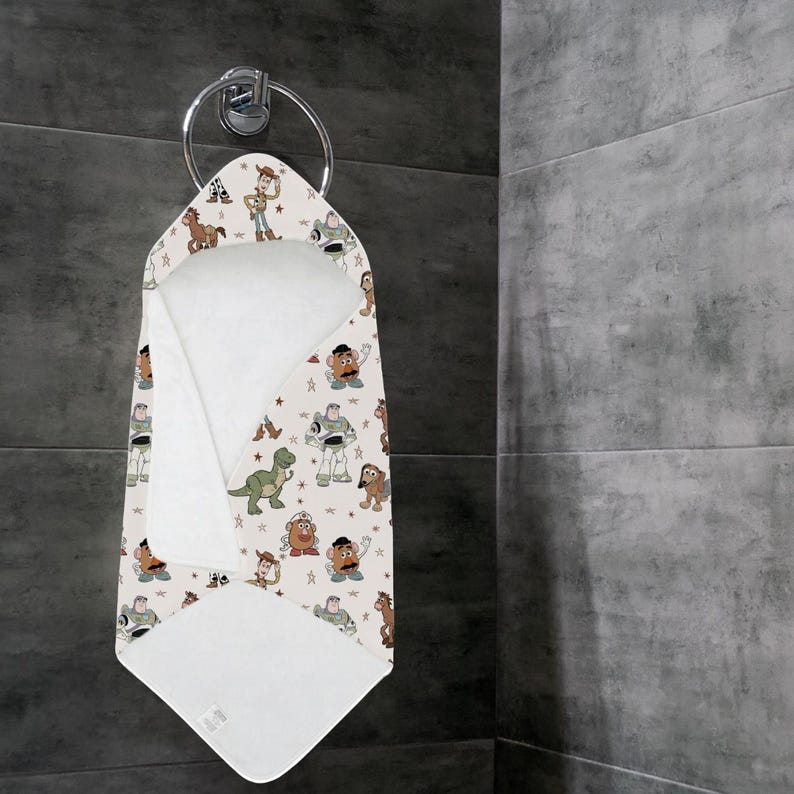 May include: A hooded baby towel with a pattern of cartoon characters, including a cowboy, a space ranger, and a green dinosaur, hangs on a silver towel ring. The towel is white on the inside and has a beige background.