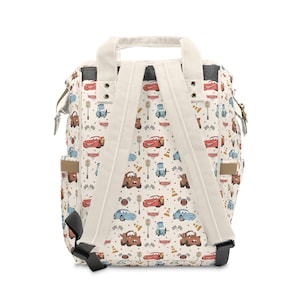 Puede incluir: Una mochila de color crema con un patr&oacute;n repetitivo de coches de carreras, sem&aacute;foros y banderas a cuadros. La bolsa incluye dos asas superiores, correas ajustables para los hombros y bolsillos laterales con detalles de herrajes met&aacute;licos.