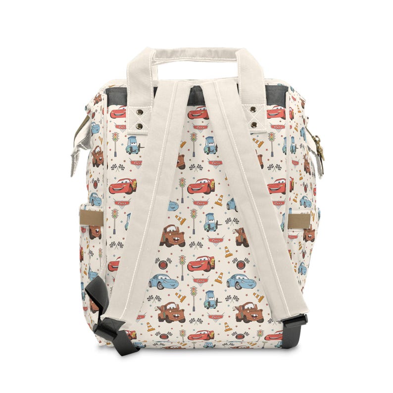 Puede incluir: Una mochila de color crema con un patr&oacute;n repetitivo de coches de carreras, sem&aacute;foros y banderas a cuadros. La bolsa incluye dos asas superiores, correas ajustables para los hombros y bolsillos laterales con detalles de herrajes met&aacute;licos.