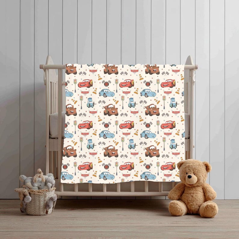 May include: A cream-coloured baby blanket featuring a repeating pattern of cartoon race cars, including red, blue, and brown vehicles, along with orange traffic cones, chequered flags, and small red banners on a light background.