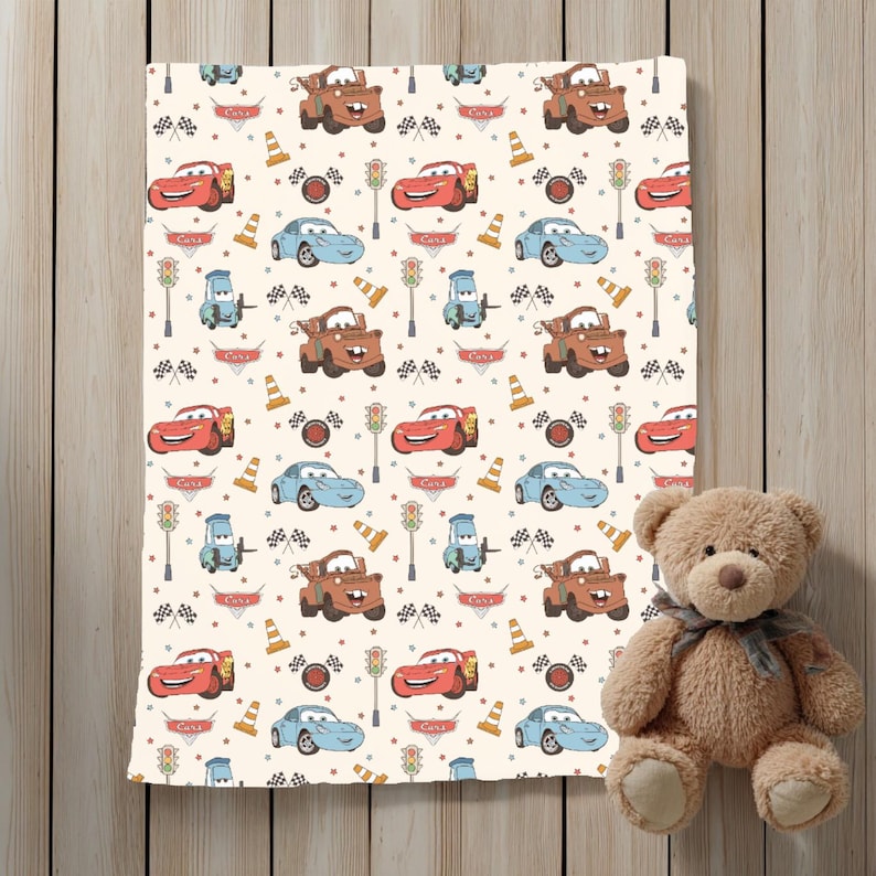 May include: A cream-coloured blanket featuring a repeating pattern of characters from the movie Cars, including red race cars, brown tow trucks, and blue sports cars, interspersed with traffic lights, orange cones, and checkered racing flags.