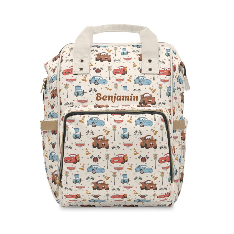 Puede incluir: Una mochila de color crema con un patr&oacute;n repetitivo de coches de dibujos animados, sem&aacute;foros y banderas a cuadros. El nombre Benjamin est&aacute; impreso en texto marr&oacute;n en la parte delantera. La bolsa incluye un asa superior, un bolsillo frontal y laterales.