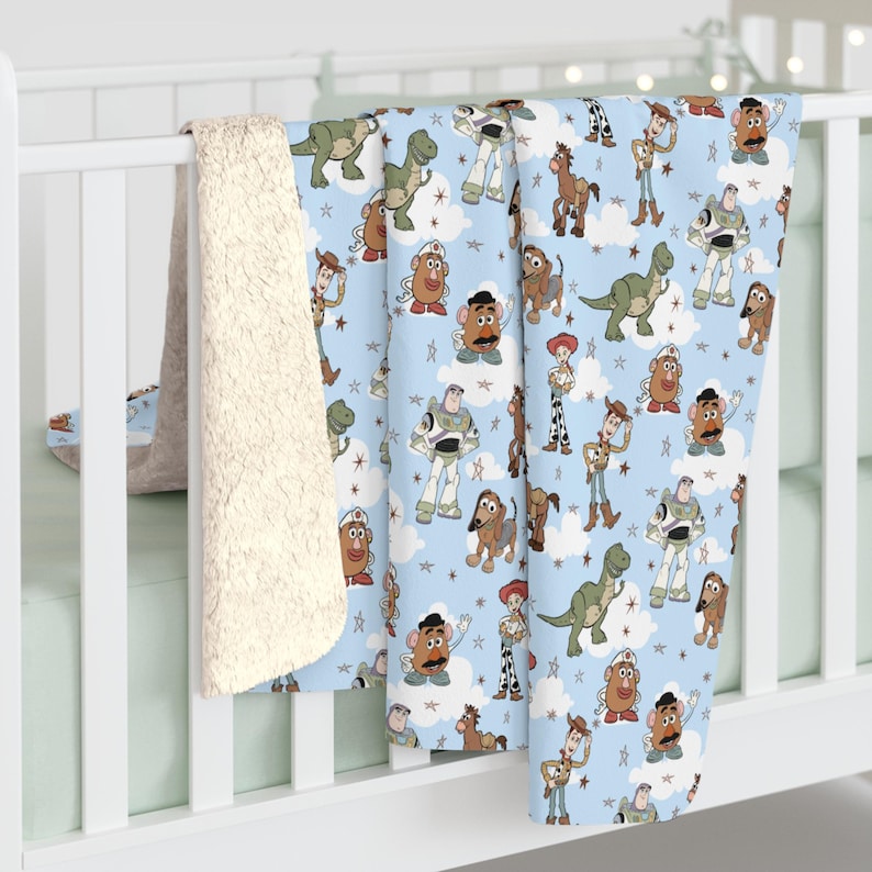 K&ouml;nnte beinhalten: Eine hellblaue Fleecedecke mit einem sich wiederholenden Muster aus Toy Story-Figuren wie Woody, Buzz Lightyear, Rex, Mr. Potato Head, Jessie, Bullseye und Slinky Dog, durchsetzt mit wei&szlig;en Wolken und kleinen Sternen auf einem weichen Stoff.