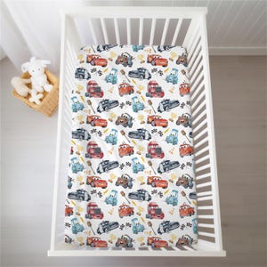 May include: White crib sheet with a repeating pattern of cartoon race cars in red, blue, black, and brown. The design includes checkered flags, trophies, and other racing-themed elements. The sheet is inside a white crib.