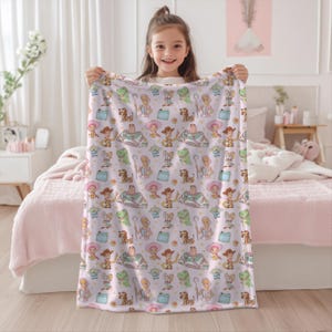May include: A light pink blanket with a repeating pattern of Toy Story characters. The blanket features characters such as Woody, Buzz Lightyear, and Rex, in a playful design. The blanket is held up by a young girl.