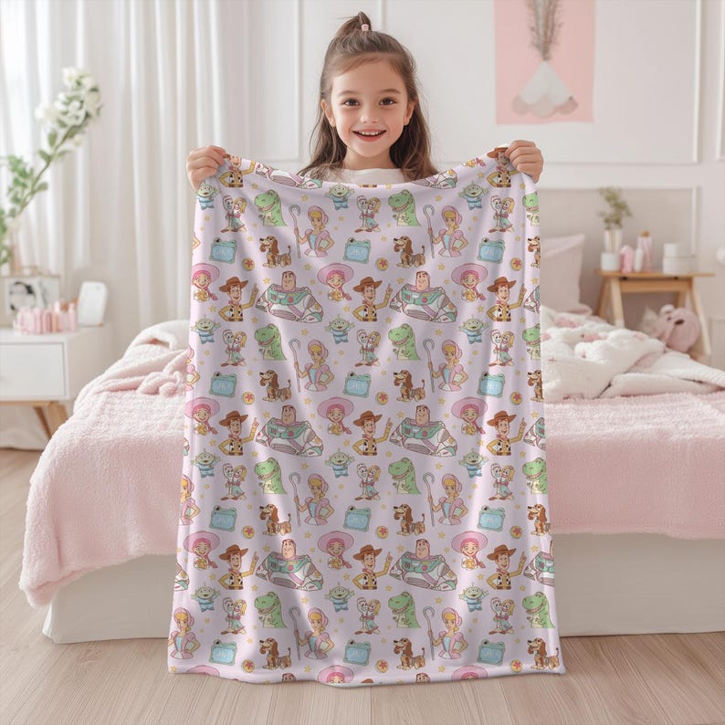 May include: A light pink blanket with a repeating pattern of Toy Story characters. The blanket features characters such as Woody, Buzz Lightyear, and Rex, in a playful design. The blanket is held up by a young girl.