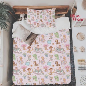 May include: A bedding set featuring a repeating pattern of cartoon characters including a space ranger, a cowboy, a shepherdess, a dinosaur, and an alien on a light pink background. The set includes a patterned pillowcase and a matching comforter.