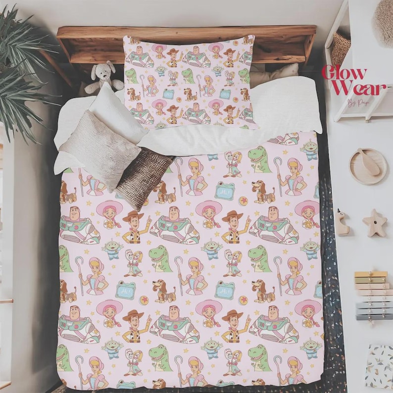 May include: A bedding set featuring a repeating pattern of cartoon characters including a space ranger, a cowboy, a shepherdess, a dinosaur, and an alien on a light pink background. The set includes a patterned pillowcase and a matching comforter.