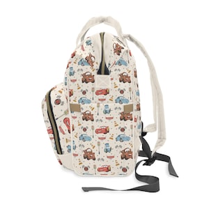 Puede incluir: Una mochila de color crema con un estampado de coches de carreras de dibujos animados, conos de tr&aacute;fico y banderas a cuadros. La bolsa incluye un asa superior, correas ajustables para los hombros, un bolsillo frontal y un compartimento principal.