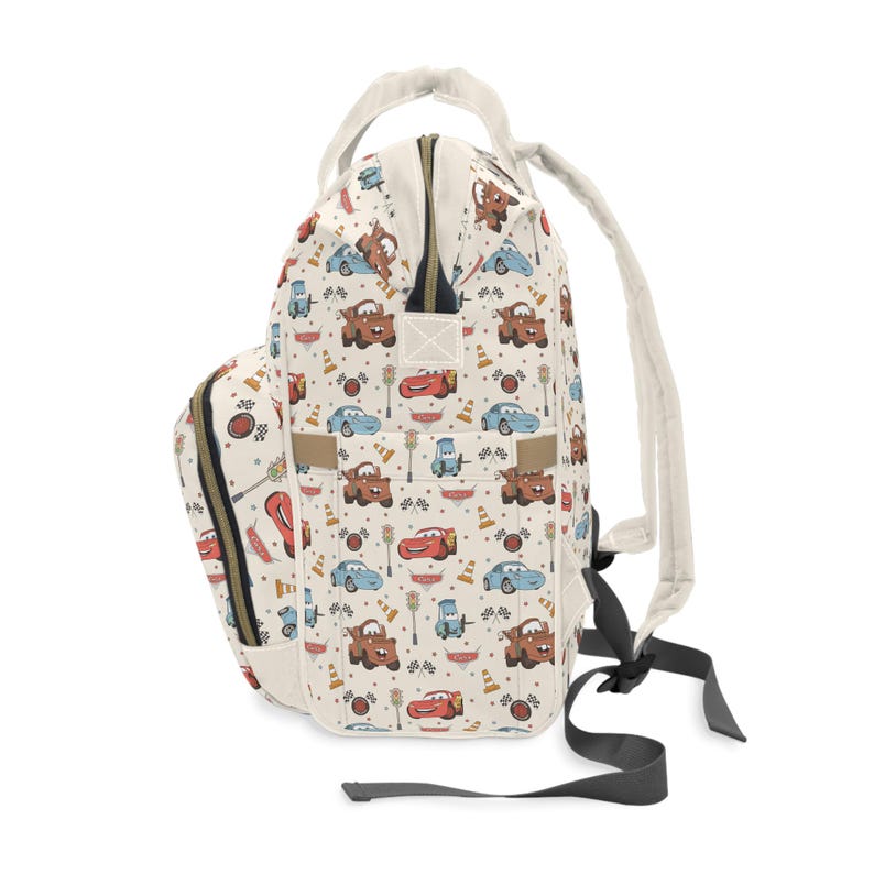 Puede incluir: Una mochila de color crema con un estampado de coches de carreras de dibujos animados, conos de tr&aacute;fico y banderas a cuadros. La bolsa incluye un asa superior, correas ajustables para los hombros, un bolsillo frontal y un compartimento principal.