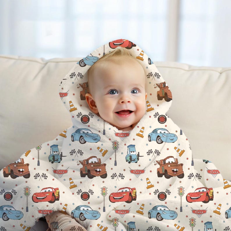 May include: A hooded baby blanket featuring a repeating pattern of characters from the movie Cars, including red and blue race cars, brown tow trucks, traffic cones, traffic lights, and checkered flags on a cream-coloured background.