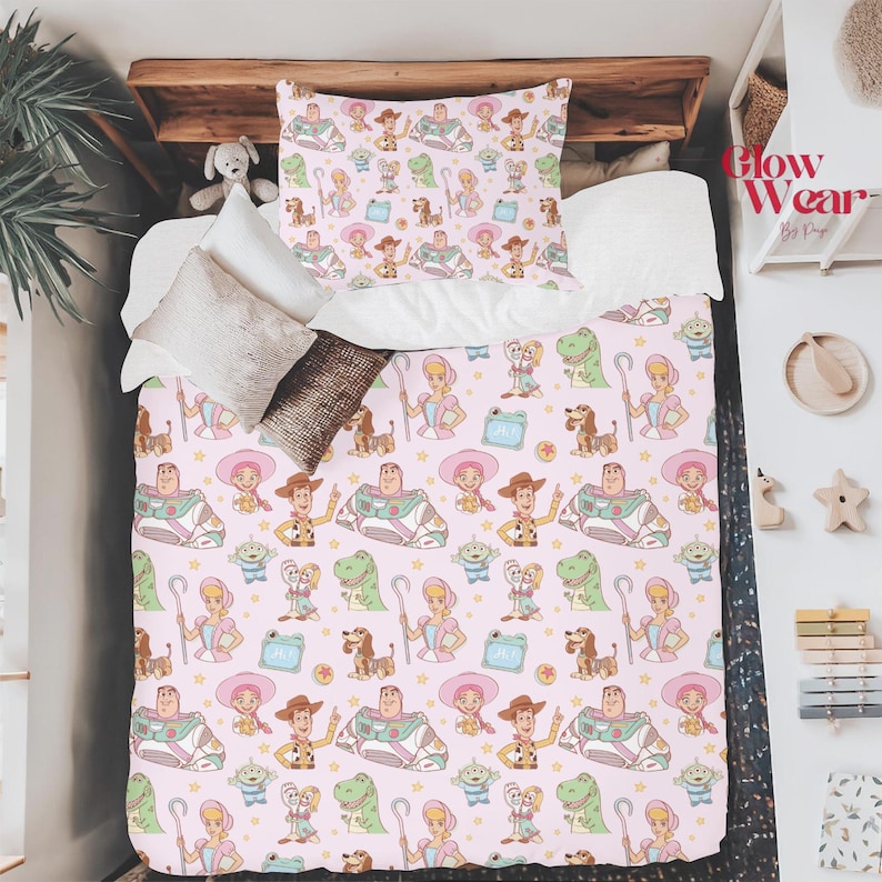May include: Pink bedding set featuring a repeating pattern of cartoon characters, including a cowboy, space ranger, dinosaur, and other toys. The set includes a pillowcase and a comforter, perfect for a child's bedroom.