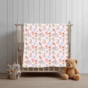 May include: A pink baby blanket featuring a repeating pattern of cartoon characters. The blanket is draped over a white cot. A brown teddy bear sits on the floor to the right of the cot, and a basket of soft toys is on the floor to the left.