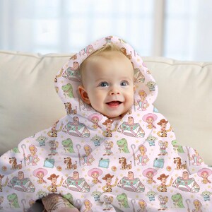 May include: A baby wearing a light pink hooded towel with a repeating pattern of Toy Story characters. The towel has a hood and is draped over the baby's shoulders. The baby is smiling.