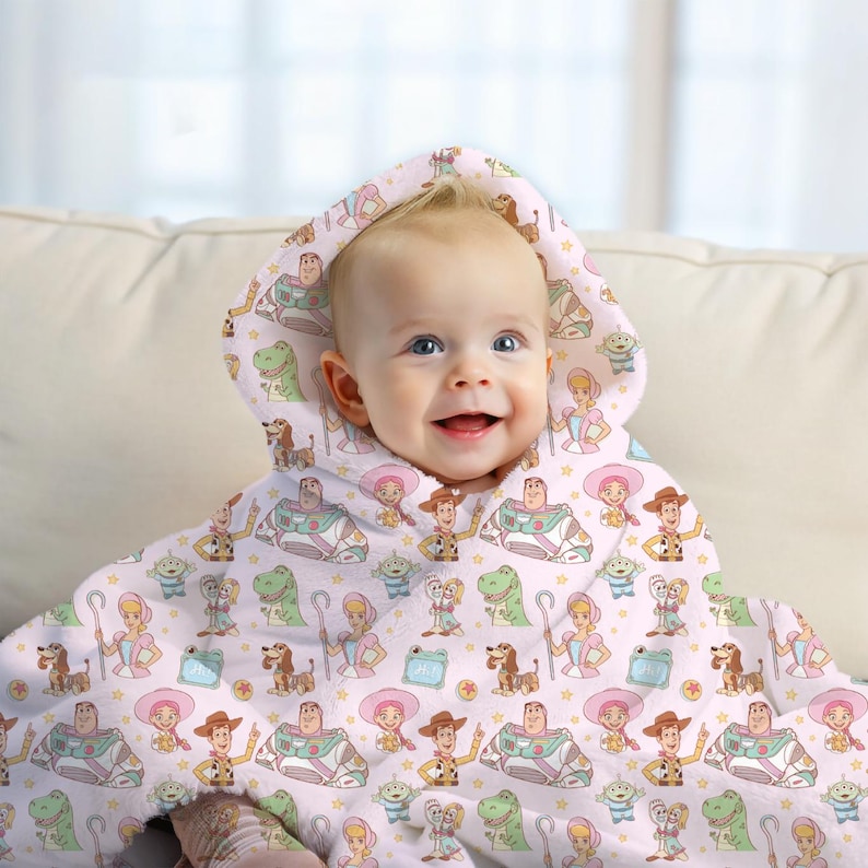 May include: A baby wearing a light pink hooded towel with a repeating pattern of Toy Story characters. The towel has a hood and is draped over the baby's shoulders. The baby is smiling.