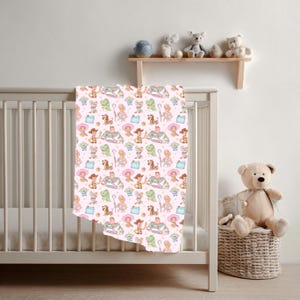 May include: A baby blanket featuring a repeating pattern of cartoon characters in various colours, including green, pink, and yellow. The blanket is draped over a white cot. A teddy bear sits in a wicker basket.